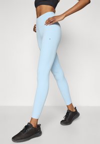 Hele sinised kõrge vöökohaga retuusid, millel on sile tekstuur ja peen logo detail, kombineeritud mustade võistlussidemetega võrgumaterjali ja kummipõhjaga spordijalatsitega.