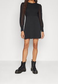 Schwarzes Kleid mit langen, durchsichtigen Puffärmeln und einem gerippten Oberteil; kombiniert mit schwarzen Schnürstiefeln im Combat-Stil mit einer dicken Sohle.