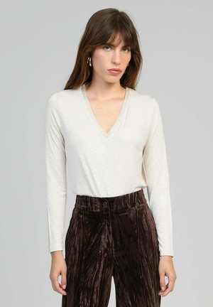 Une femme aux longs cheveux bruns porte un haut à manches longues V en beige clair et un pantalon en velours marron foncé taille haute, le tout sur un fond uni.