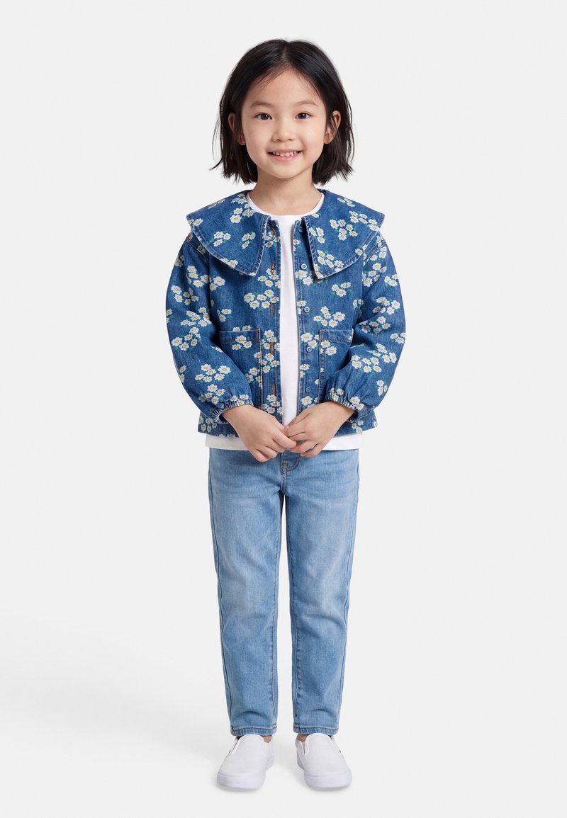 Jeune fille souriante portant une veste en jean à motifs floraux, une chemise blanche, un jean bleu clair et des chaussures blanches sans lacets, debout devant un fond uni.