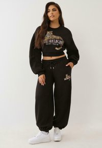 Sieviete, tērpusies melnā īsajā sporta jakā un atbilstošajās sporta biksēs ar leoparda un "True Religion Brand Jeans" logotipu, baltās kedās, stāvot.