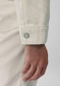 Crèmekleurige denim shirtmanchet met een gestructureerde afwerking, voorzien van een ronde metalen knoop met de opdruk "SOLVER'S" en subtiele stikdetails.