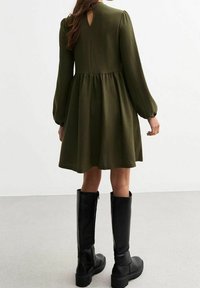 Donna in un abito lungo a maniche lunghe verde scuro con un'apertura a chiave sul retro, abbinato a stivali neri fino al ginocchio, in piedi su un pavimento grigio.