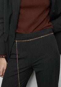 Donna che indossa un completo scuro a righe e un maglione marrone, con una sottile cintura in oro che pende dalla vita.