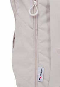 Hellgrauer Rucksack mit gepolsterten Abschnitten, einem zentralen Reißverschluss, Seitentaschen und einem Markenetikett mit roten und blauen Punkten. Glatte Stoffstruktur.