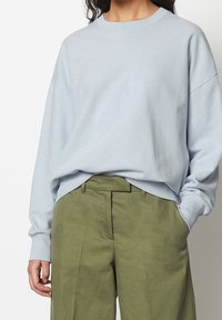 Donna che indossa un ampio felpa leggera di colore blu chiaro infilata in pantaloni a gamba larga verde oliva a vita alta, con una mano in tasca.