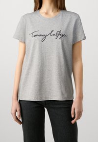 Camiseta de algodón gris de manga corta con un logo en script negro "Tommy Hilfiger" en el pecho. Diseño clásico de cuello redondo.
