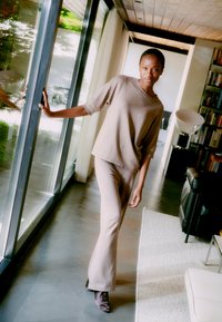 Beige gestrickter Zweiteiler, der ein lässig geschnittenes Kurzarmoberteil und ausgestellte Hosen umfasst. Weiche Textur, minimalistisches Design und sanfte Farbtonvariationen.