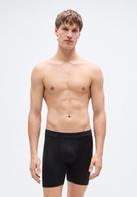 Μαύρα βαμβακερά μποξεράκια με λεία υφή, ελαστική μέση και εφαρμοστή γραμμή, συνδυασμένα με γυμνό κορμό σε απλό φόντο.
