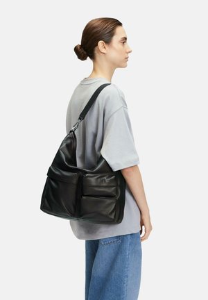 Schwarze Leder-Schultertasche mit einer lässigen Form, zwei Vordertaschen und einem breiten Trageriemen, getragen von einem Model in einem grauen, oversized Shirt.