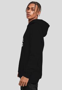 Jeune homme avec des cheveux tressés portant un sweat à capuche noir, se tenant de profil contre un fond gris clair uni.