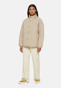 Beige gewatteerde jas met een hoge kraag, drukkers en zijzakken, gedragen met crèmekleurige broeken en bruine sneakers met witte zolen.