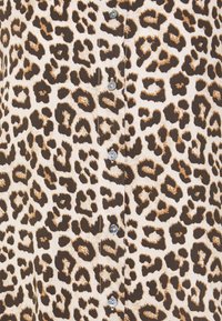 Løveprint stof i beige med brune og sorte pletter. Har sølvknapper langs fronten og en blød, tekstureret overflade.