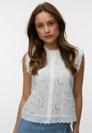 Nainen, jolla on pitkät ruskeat hiukset, pukeutuneena valkoiseen kirjailtuun hihattomaan puseroon, jossa on aaltoilevat reunat, ja sinisiin farkkuihin, yksinkertaista taustaa vasten.