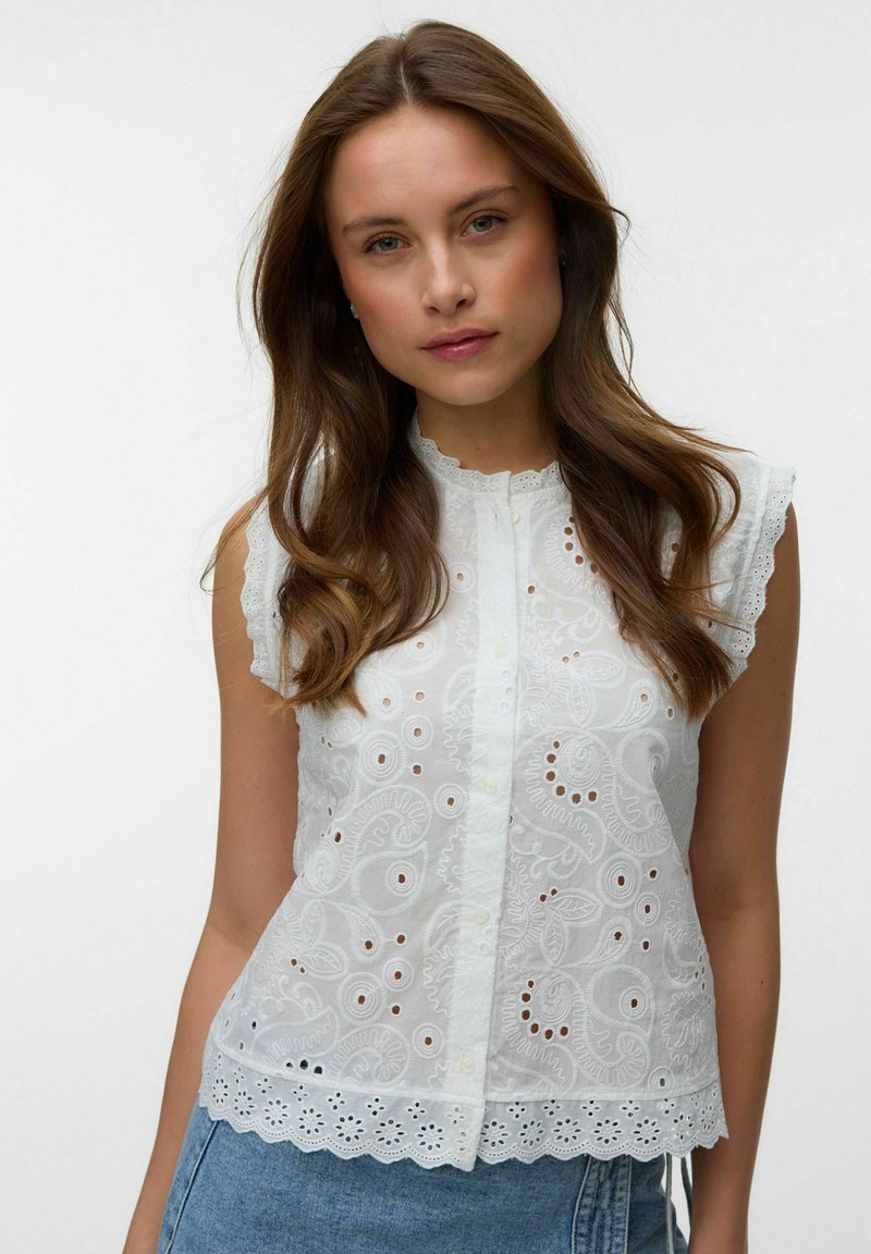 Femme aux longs cheveux bruns portant un chemisier blanc brodé sans manches avec des bords festonnés et un jean bleu sur un fond uni.