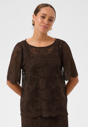 Femme aux cheveux châtain clair en chignon bas portant des boucles d'oreilles créoles dorées et un haut en dentelle florale marron foncé à manches courtes, debout sur un fond uni.