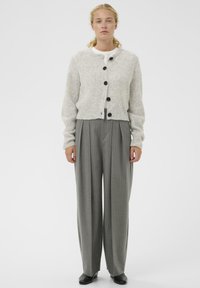 Moteris stovi vilkėdama šviesiai pilką mygtukais užsegamą megztinį, baltą marškinėlius, pilkus dryžuotus plačius kelnes ir juodus batus prieš baltą fono.