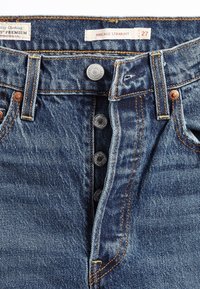 Gros plan sur un jean denim bleu avec une braguette à boutons ouverte, des boutons métalliques visibles, des passants de ceinture et des étiquettes à l’intérieur de la ceinture.