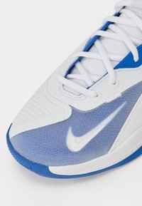 Chaussure de sport Nike blanche et bleue, avec une texture en mesh, des accents synthétiques lisses et une zone de orteil à motif pour une meilleure adhérence.