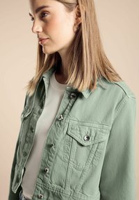 Veste en denim verte au design court, avec des détails en boutons, deux poches à rabat à l'avant et une finition texturée, portée sur une chemise blanche.