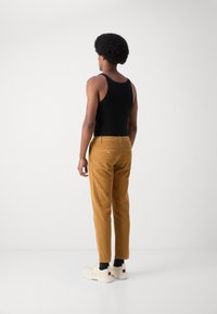 Tan corduroy broek met slim fit en verticale strepen, gecombineerd met een zwart tanktop en witte sneakers. Achteraanzicht, minimalistische achtergrond.