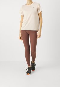 Ljusrosa tränings t-shirt med en liten logotyp, matchad med mörkbruna leggings och svarta sneakers med orange detaljer.