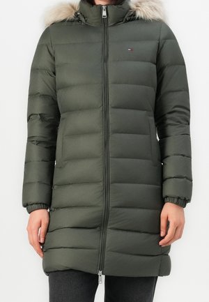Femme portant un long manteau matelassé vert foncé avec fermeture éclair à l'avant et capuche bordée de fourrure, debout devant un fond uni.