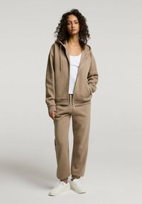 Beżowy hoodie na zamek z kieszeniami z przodu, w zestawie ze spodniami dresowymi w tym samym kolorze. Biała koszulka na ramiączkach pod spodem i białe sneakersy. Gładka bawełniana tekstura.