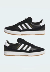Zapatillas deportivas negras con parte superior de ante y cuero sintético, tres franjas blancas, suela de goma y detalles en dorado.