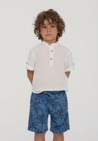 Camicia bianca in lino a maniche corte con colletto alla mandarino e bottoni marroni, abbinata a pantaloni corti blu con stampa di palme.