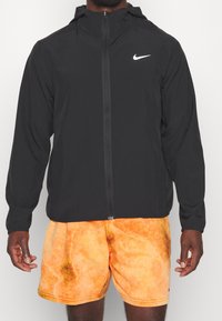 Man wearing black Nike zip-up jacket and orange patterned shorts, standing against white background, arms relaxed at sides.