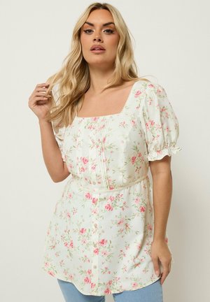 Blond kvinde iført en hvid blomsterknap-frem-ballonærme-bluse, stående mod en ensfarvet lys baggrund.