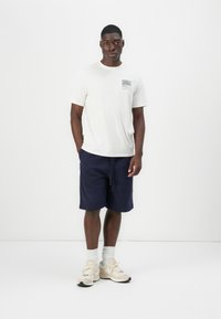 Kremfarget T-skjorte med kort erm og grafikk foran, kombinert med marineblå shorts. Modellen har på seg kremfargede joggesko med lyse sokker. Enkel, uformell antrekk.