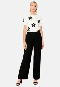 Top bianco lavorato a maglia con applicazioni di fiori neri, abbinato a pantaloni a gamba larga neri a vita alta. Modella che indossa tacchi neri, braccia rilassate.