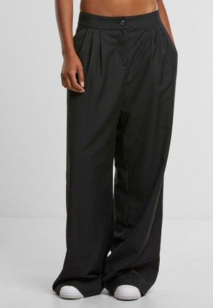 Femeie purtând pantaloni negri largi cu talie înaltă, buzunare laterale și adidași albi, stând în picioare în fața unui fundal simplu.