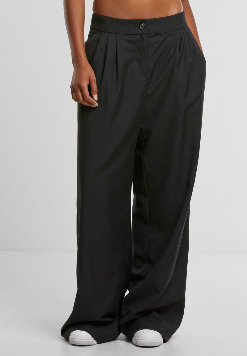 Woman wearing loose black high-waist wide-leg pants with side pockets and white sneakers, standing against a plain background.