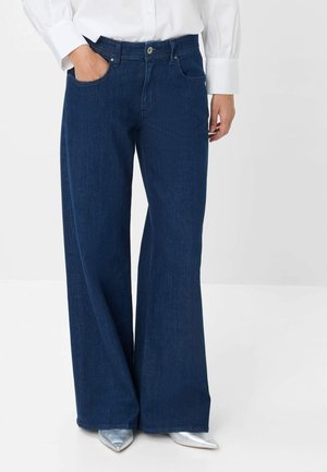 Henkilö pukeutuneena tummansinisiin leveälahkeisiin farkkuihin, valkoiseen pitkähihaseen paitaan ja hopeisiin kärjellisiin kenkiin yksinkertaista taustaa vasten.