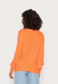 Oranje gebreide trui met een losse pasvorm, voorzien van lange mouwen, een gestructureerd patroon en een ribgebreide zoom. Gekleed met licht gewassen jeans.