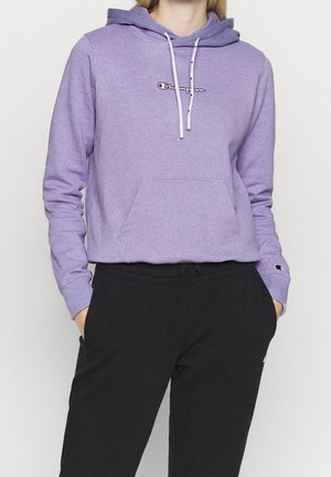Vrouw die een lavendelkleurige Champion hoodie met witte trekkoorden en een zwarte broek draagt, staand met handen in de zakken tegen een effen achtergrond.