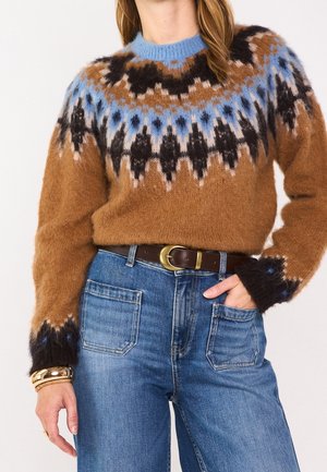 Femme portant un pull en maille à motifs marron, bleu et noir, rentré dans un jean taille haute bleu avec une ceinture marron et un bracelet doré.