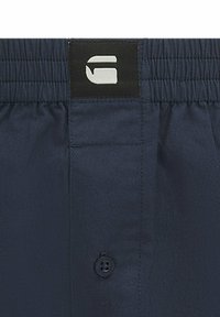 Navyfarbene Shorts mit elastischem Bund, versehen mit einem schwarzen Patch, auf dem ein stilisiertes weißes "G"-Logo und ein einzelner schwarzer Knopf darunter abgebildet sind.