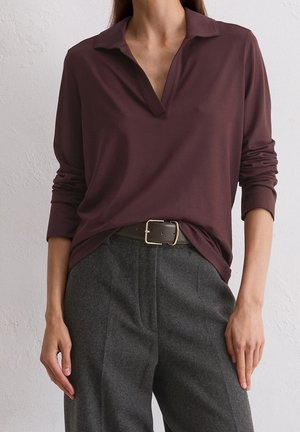 Frau trägt ein dunkelviolettes langärmeliges Oberteil, das in graue hoch taillierte Hosen mit einem dunkelbraunen Gürtel und goldener Schnalle gesteckt ist.