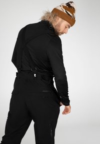 Salopette da sci nera con bretelle regolabili, tasche funzionali e branding discreto, abbinata a un cappellino di maglia a righe marroni e bianche.