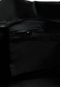 Interior de una bolsa de tela negra con un bolsillo con cremallera visible. La textura es suave con un patrón uniforme. La cremallera es de tono plateado.