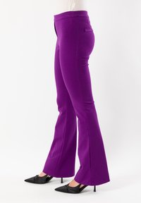 Pantaloni a zampa d'elefante viola realizzati in tessuto elasticizzato con una texture liscia. Presentano una tasca frontale e una vita aderente. Indossati con tacchi neri.