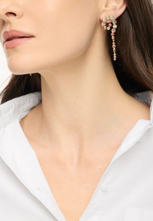 Kvinde iført en guldring formet som et spørgsmålstegn med pink og grønne ædelsten, parret med en hvid skjorte med krave.