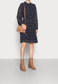 Mekko, jossa on laivaston sininen kukkakuosi, kerätty vyötärö, pitkät hihat hupullisilla hihoilla ja kevyt kangas. Vaaleanruskea olkalaukku ja nilkkurit täydentävät lookin.