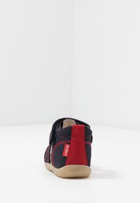 Zapato infantil en tonos azul marino y rojo con parte superior de ante y textil, correa de velcro y suela de goma blanda. Presenta costuras rojas y una marca distintiva.