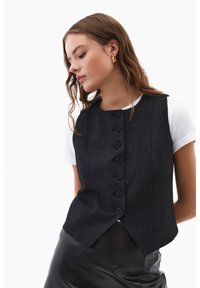 Sort vest med fine striber, fem knapper, høj halsudskæring og skrå kant, lagdelt over en hvid kortærmet skjorte. Glat stoftekstur.