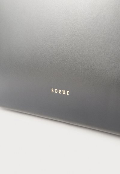 Κοντινό πλάνο λείου γκρι δερμάτινου υλικού με μικρό, χρυσό φολιδωτό κείμενο "soeur" τοποθετημένο στο κέντρο κοντά στο κάτω άκρο.